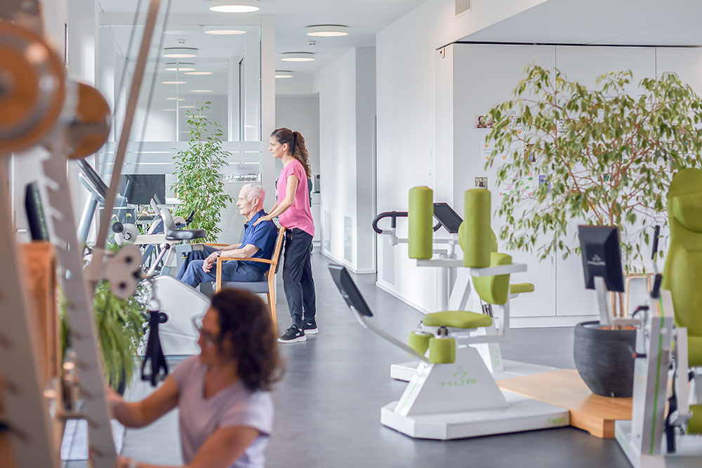 Zwei Physiotherapeutinnen unterstützen einen Bewohner bei Übungen im hellen und freundlich gestalteten Trainingsraum.
